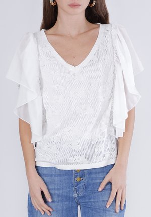 Kobieta ubrana w biały top z dekoltem w serek z motywem motyli i przezroczystymi, warstwowymi falbanianymi rękawami, w połączeniu z niebieskimi dżinsami.