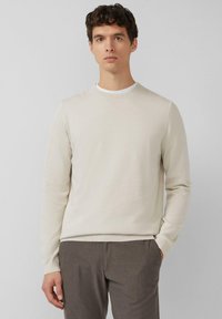 Beiger Pullover mit langen Ärmeln, geripptem Rundhalsausschnitt und strukturierter Strickmuster, getragen zu grauen gestreiften Hosen, Hände in den Taschen.