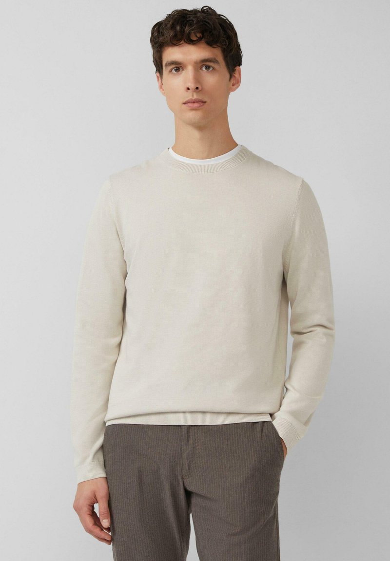 Beiger Pullover mit langen Ärmeln, geripptem Rundhalsausschnitt und strukturierter Strickmuster, getragen zu grauen gestreiften Hosen, Hände in den Taschen.