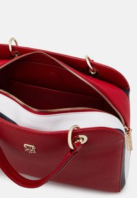 Sac en cuir rouge avec une surface texturée, des anses en or et un intérieur contrastant blanc et marine. Comprend des fermetures à glissière doubles et des poches intérieures.
