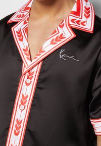 Black satin shirt with white and red patterned collar and button line, short sleeves, and white embroidered signature on the chest.