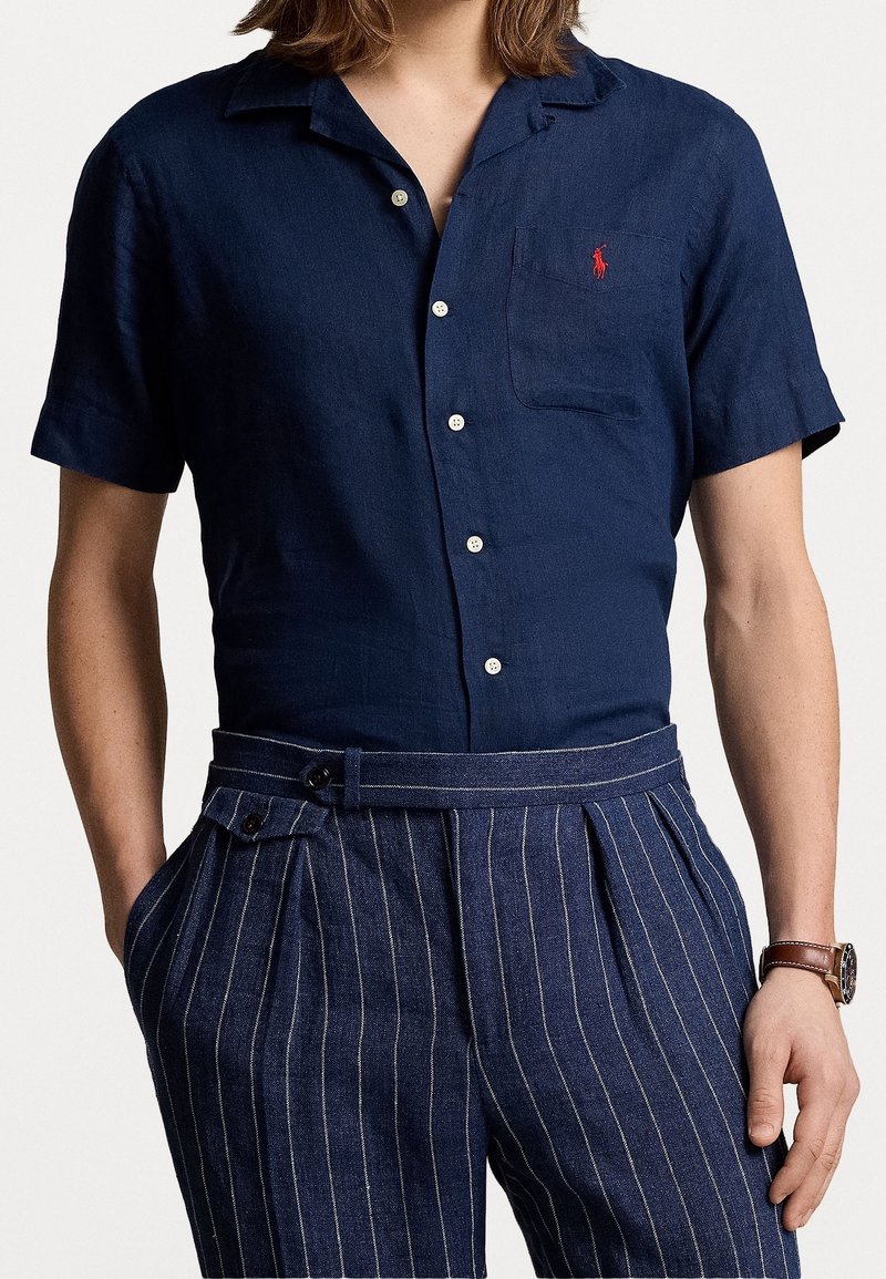 Navy linen short-sleeve shirt with a collar, button closure, and a chest pocket. Paired with navy pinstriped pleated trousers.