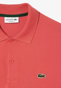 Korallfärgad polotröja med strukturerad stickning, med en grön detalj vid kragen, två knappar och en broderad krokodil-logotyp på bröstet.