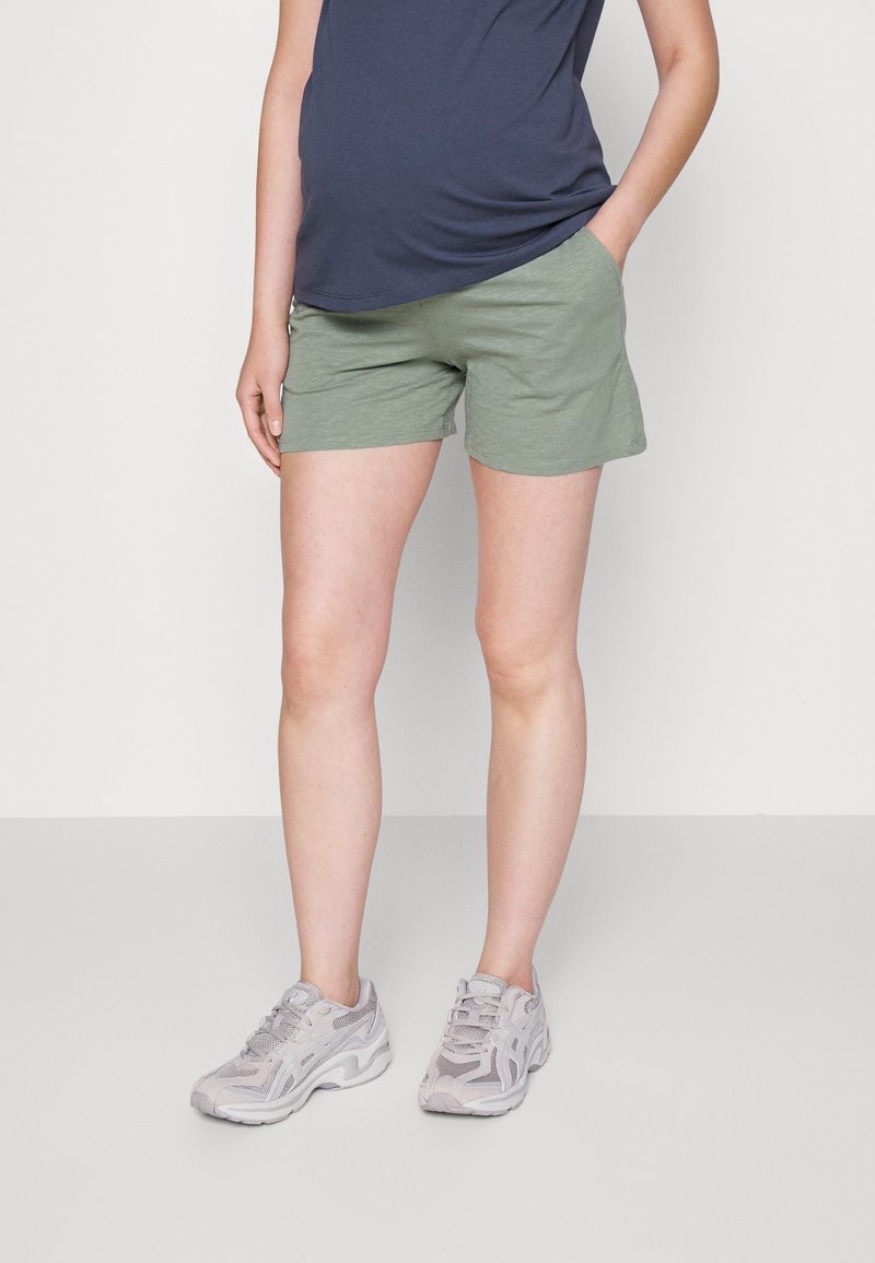 Personne portant des baskets gris clair, un short vert olive et un haut bleu marine, une main dans la poche, debout sur un sol gris.