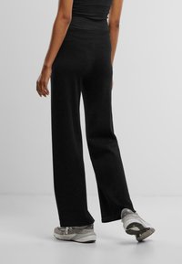 Schwarze, weite Hosen aus weichem, dehnbarem Material. Nahtloser Bund; kombiniert mit hellgrauen Sportschuhen mit dunklen Akzenten.