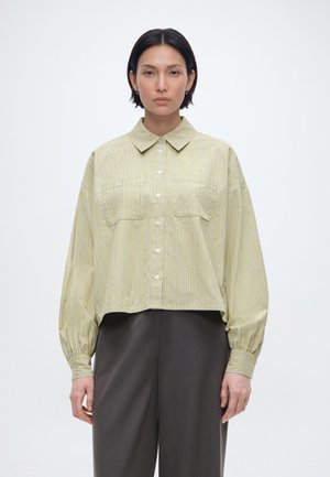 Žena s rovnými čiernymi vlasmi, oblečená v svetložltej pruhovanej košeli s veľkými manžetami a tmavosivých nohaviciach, čelom k fotoaparátu.