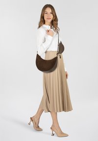 Brown suede crescent-shaped handbag with gold hardware and an adjustable strap, worn on the shoulder with a beige pleated skirt.