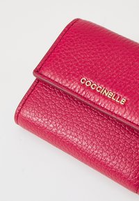 Rožinė odinė piniginė su akmeniniu tekstūru, su auksiniu "Coccinelle" logotipu priekyje ir sulankstomu dangteliu.