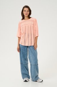 Blusa de color coral con detalles de ojales y mangas anchas, combinada con pantalones de mezclilla azules holgados y zapatillas blancas. Conjunto simple y casual.