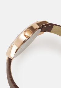 Roségoldene Uhr mit einem polierten Metallgehäuse, großem runden Ziffernblatt, braunem Lederarmband und einer strukturierten Krone an der Seite.