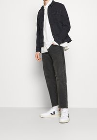 Giacca di jeans nera, camicia bianca con bottoni, jeans grigio scuro, sneaker bianche con dettagli. L'outfit è casual e moderno nello stile e nei colori.