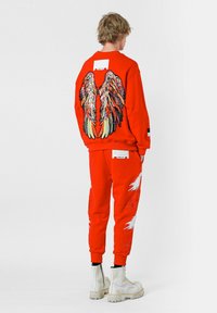 Sudadera roja con diseño de alas de lentejuelas multicolores en la parte posterior, combinada con pantalones a juego con detalles blancos y una etiqueta parche. Botas blancas.