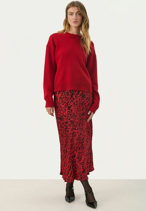 Roter Strickpullover mit langen Ärmeln und rundem Halsausschnitt, kombiniert mit einem roten Midirock im Leopardenmuster. Schwarze Ankle-Strap-Heels runden den Look ab.