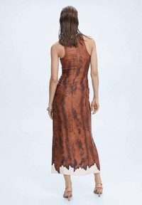 Frau mit nassen Haaren, die ein ärmelloses, braunes Batik-Midikleid und nudefarbene, hochhackige Sandalen trägt, stehend vor einem schlichten, hellen Hintergrund.