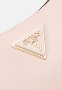 Lyserød blush-farvet taske i kunstlæder med en trekantet emblem i guldtonet metal med teksten "GUESS". Glat tekstur og enkel design, der fremhæver brandingsdetaljen.