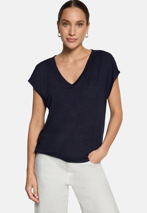 Femme portant un haut bleu marine à col en V et manches courtes, ainsi qu'un pantalon blanc, debout avec une main dans la poche devant un fond uni.