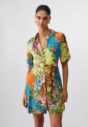 Femme portant une robe courte boutonnée à imprimé floral multicolore et animal, avec une ceinture nouée et des bijoux dorés, posant devant un fond uni.