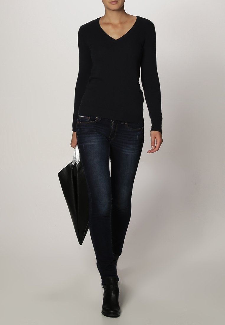 Black long-sleeve top with a v-neck, paired with dark blue skinny jeans, and black ankle boots. Model holds a large black tote bag.