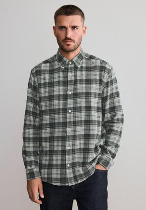 Man with short hair and beard wearing a gray and black plaid button-up shirt, standing with one hand in pocket against light background.