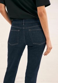 Jeans de mezclilla oscura con corte ajustado, que cuentan con bolsillos traseros, costuras en contraste y una textura suave. Combinados con una camisa negra.