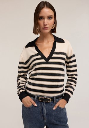 Femme portant un pull V en col noir et beige à rayures, un jean bleu, une ceinture noire avec une boucle en or et de grandes boucles d'oreilles en or.