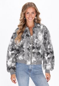 Donna con lunghi capelli biondi mossi che sorride, indossando una giacca con zip grigio e bianco a trama e jeans azzurri chiari, posando con le mani rilassate.