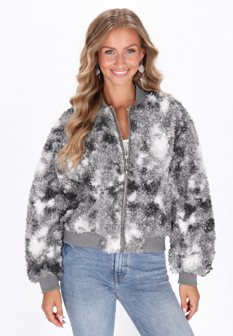 Donna con lunghi capelli biondi mossi che sorride, indossando una giacca con zip grigio e bianco a trama e jeans azzurri chiari, posando con le mani rilassate.