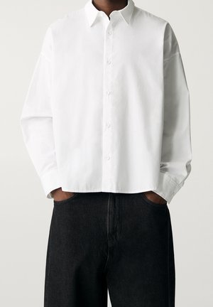 Person wearing a loose white button-up shirt with long sleeves tucked casually into high-waisted black pants, hands in pockets.