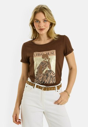 Mujer rubia vistiendo una camiseta gráfica marrón con el diseño "Urban Muse" y pantalones blancos, complementada con un cinturón marrón y una pulsera dorada.