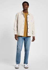 Chaqueta de tergal color beige claro con bolsillo frontal, camiseta mostaza debajo, jeans azules y zapatillas blancas con acentos en negro y gris.