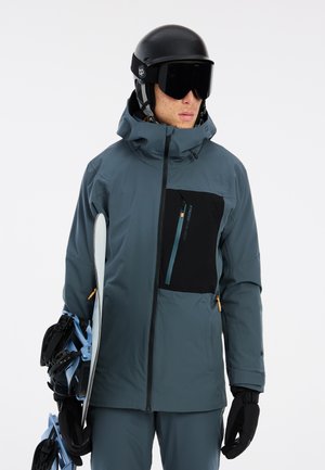 Chaqueta de nieve en color teal para hombre con un bolsillo con cremallera negra, capucha ajustable y cremalleras azules en contraste. Los accesorios incluyen un casco negro y una tabla de snow.