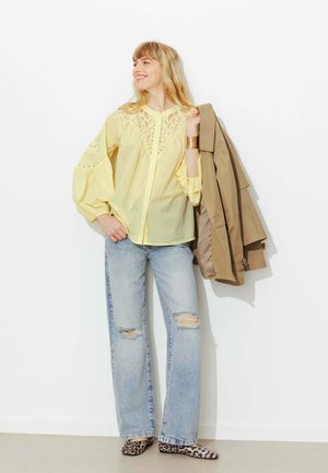 Femme vêtue d'un chemisier jaune en dentelle et d'un jean déchiré, tenant un manteau beige sur l'épaule, portant des chaussures à imprimé léopard et un bracelet manchette en or.