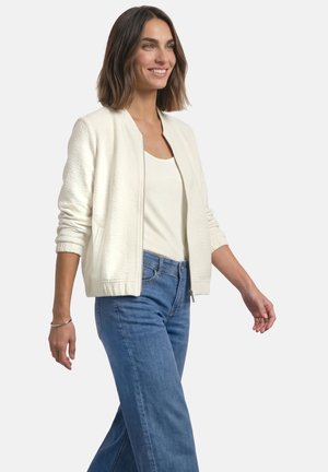 Kvinde smilende og gående iført en hvid tekstureret lynlæserjakke, hvid top, blå jeans og et sølvarmbånd på venstre håndled.