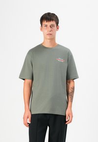 Olijfgroene katoenen t-shirt met korte mouwen, ronde hals en een logo op de borst in rood en blauw, gecombineerd met zwarte broek.