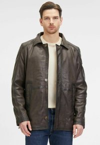 Braune Lederjacke mit Kragen, vorderem Knopfverschluss, zwei Seitentaschen und strukturiertem Finish. Getragen über einem cremefarbenen Hemd und Jeans.