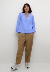 Blusa azzurro chiaro a maniche lunghe con scollatura a fiocco, dettagli arricciati e una vestibilità rilassata, abbinata a pantaloni beige e scarpe da ginnastica grigie.