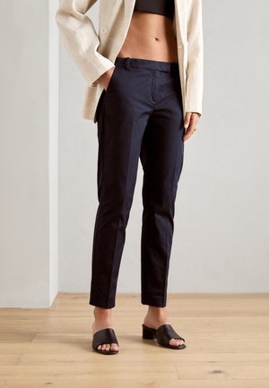 Femme portant un pantalon droit bleu marine, un blazer beige, un top court noir et des sandales à talons ouvertes noires, debout sur un sol en bois.