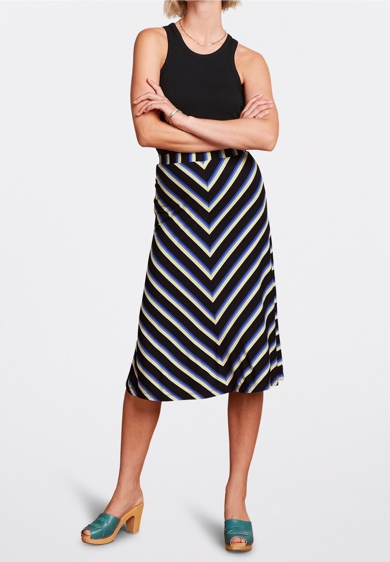 Černý žebrovaný tílko spojené s proužkovanou sukní se vzorem chevron v modré, černé a krémové barvě. Modelka má také na nohou tyrkysové dřeváky.