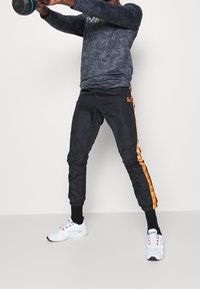 Mustat urheiluhousut oransseilla sivuraidalla, kapeneva muotoilu ja elastiset hihat. Yhdistetty pitkähihainen tumma kamotoppi ja valkoiset lenkkarit.