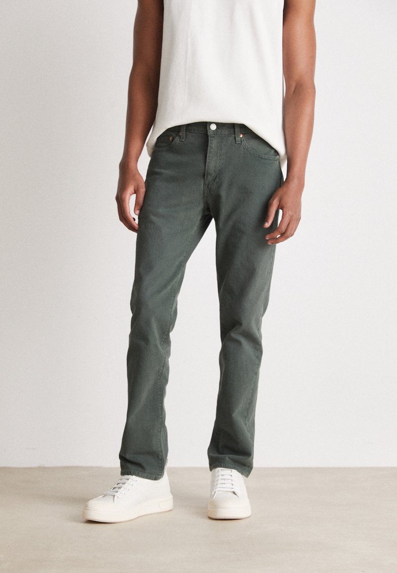 Personne portant un pantalon droit vert foncé, des baskets blanches et un haut blanc sans manches devant un fond neutre.