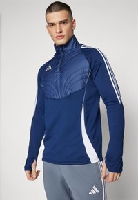 Marineblaue Sportjacke mit einem halben Reißverschluss, gepolstertem Brustbereich und weißen Seitenstreifen; glattes Material mit einer detaillierten Steppung auf der Vorderseite.