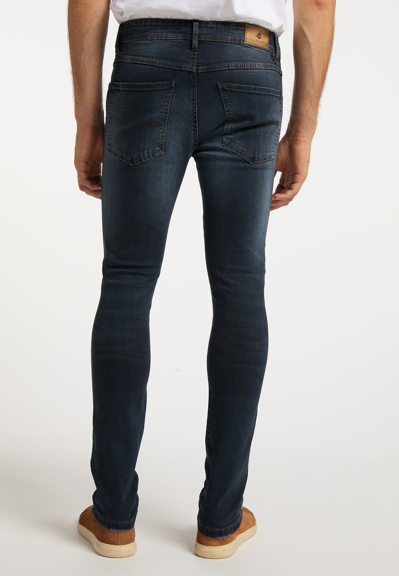Schmal geschnittene dunkelblaue Jeans mit Gesäßtaschen, minimalen Verwaschungen und einem hellbraunen Lederetikett am Bund. Füße in braunen Schuhen sichtbar.