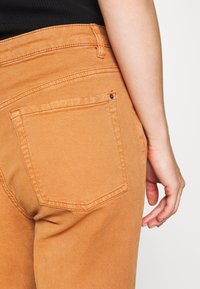 Los pantalones de mezclilla naranja cuentan con un bolsillo trasero con costuras visibles y un remache de metal. La tela tiene una textura suave y una silueta ajustada.