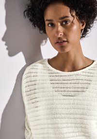 Helles cremefarbenes Strickpullover mit horizontalen Streifen. Der Pullover hat einen weiten Ausschnitt und einen lässigen Schnitt. Der Schatten des Models fällt auf eine schlichte Wand.