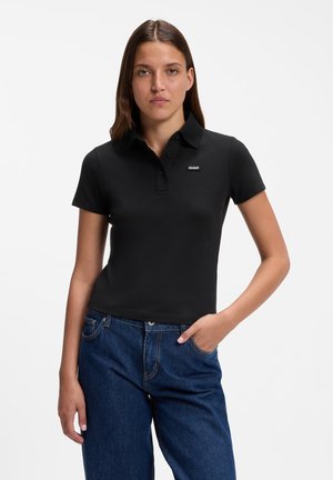Ung kvinde med glat brunt hår iført en sort kortærmet polo og blå jeans, stående med den ene hånd i lommen.