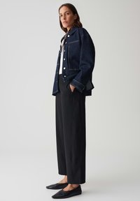 Veste en denim bleu foncé avec des coutures claires, associée à un pantalon noir à jambes larges et des ballerines noires à bout pointu. Le haut en dessous est blanc.