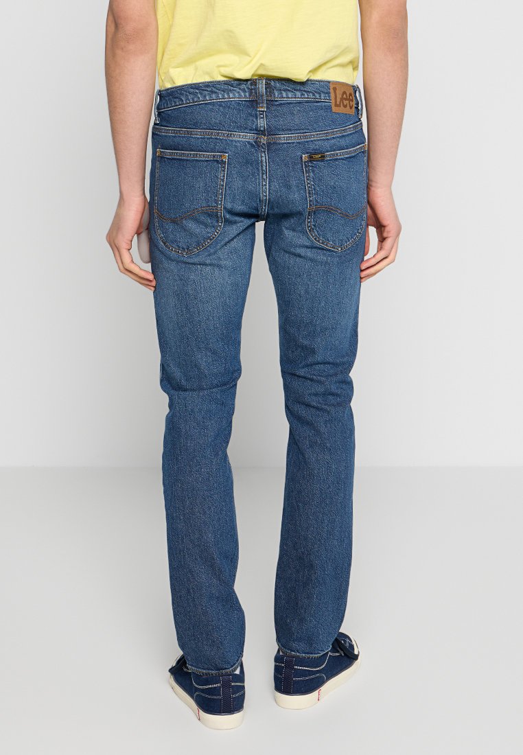 Mężczyzna w niebieskich dżinsach Lee o prostym kroju, żółtej koszulce i granatowych sneakersach stoi plecami do aparatu na jednolitym szarym tle.