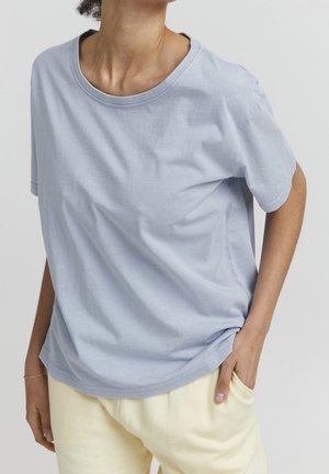 Persona con camiseta de manga corta azul claro y pantalones color crema, con una mano en el bolsillo frente a un fondo claro y sencillo.