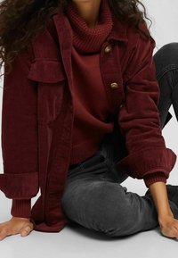 Veste en velours côtelé bourgogne avec des boutons en laiton superposée sur un pull à col roulé côtelé. Jeans noirs au toucher doux, en position assise.