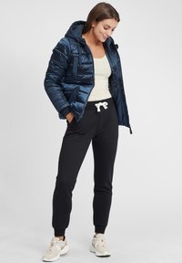 Tumši zils pūkains skapīša jacket ar kapuci un ar rāvējslēdzēju, apvienots ar gaišu topu un melnām sporta biksēm. Baltā aukliņa detaļa biksēs.